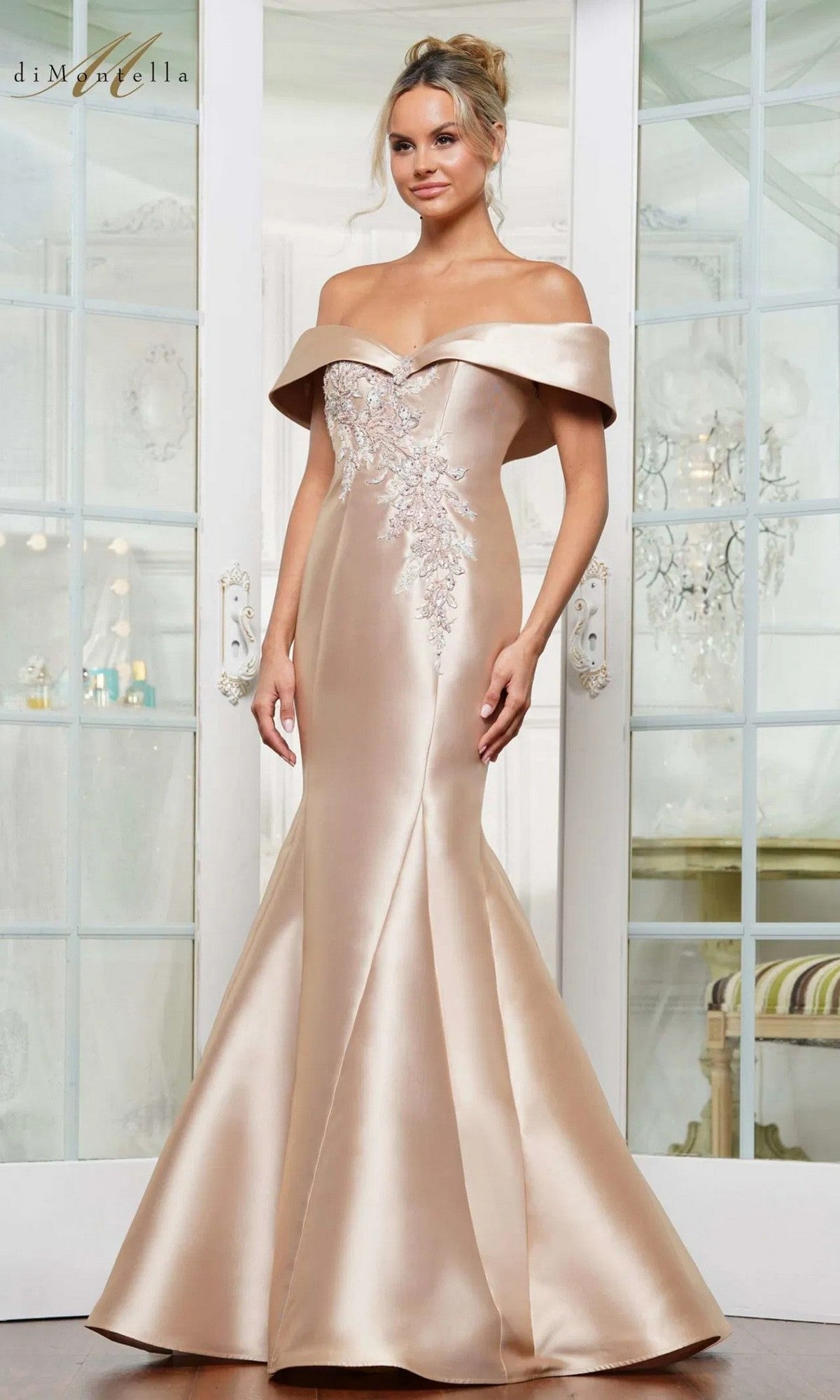A woman in the gold Long Formal Dress Rina Di Montella RD3257, featuring an off-the-shoulder neckline, beaded floral embroidery, and a mermaid silhouette, stands in front of glass-paned doors.